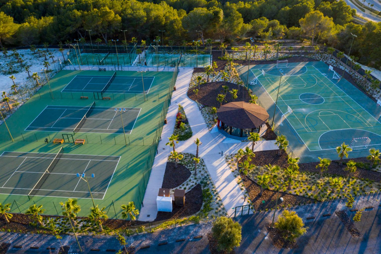 Aerial view of a recreational complex with multiple fenced tennis courts on the left and a large multiuse court on the right, surrounded by trees and a central gazebo.