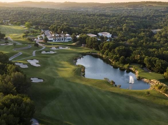 Aerial view of a lush golf course with sand traps, a winding fairway, and a central pond near a clubhouse surrounded by trees.