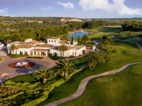 Aerial view of a white villa-like resort complex with palm trees, circular flower bed, and a nearby golf course.