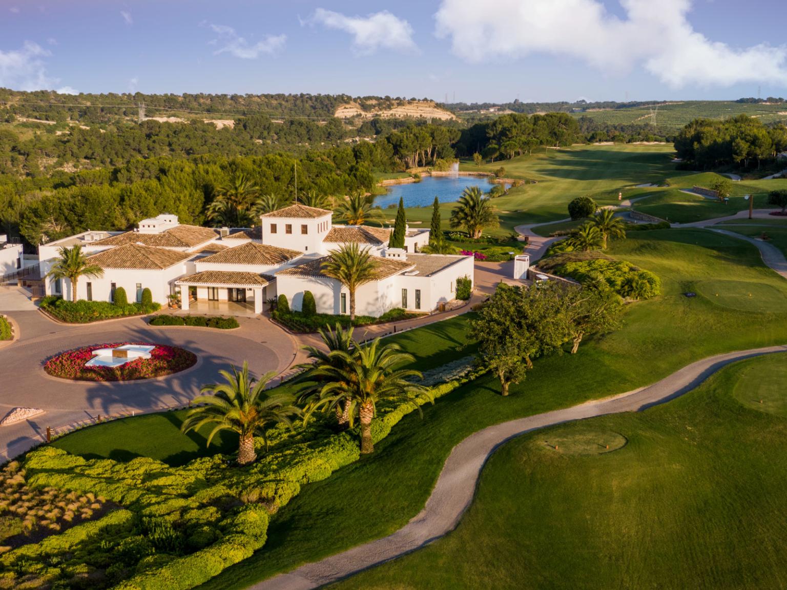 Aerial view of a white villa-like resort complex with palm trees, circular flower bed, and a nearby golf course.