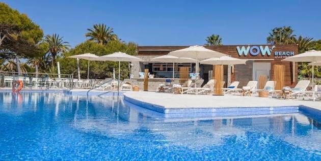 Outdoor resort pool with blue water, white lounge chairs, and palm trees; building labeled 'WOW Beach' in background.