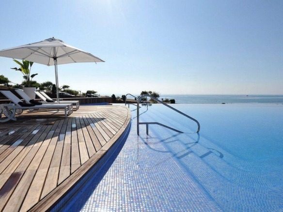 Infinity pool with a wooden deck, sun loungers, and a large white parasol overlooking the ocean sunlit scene.
