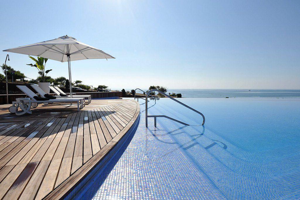 Infinity pool with a wooden deck, sun loungers, and a large white parasol overlooking the ocean sunlit scene.