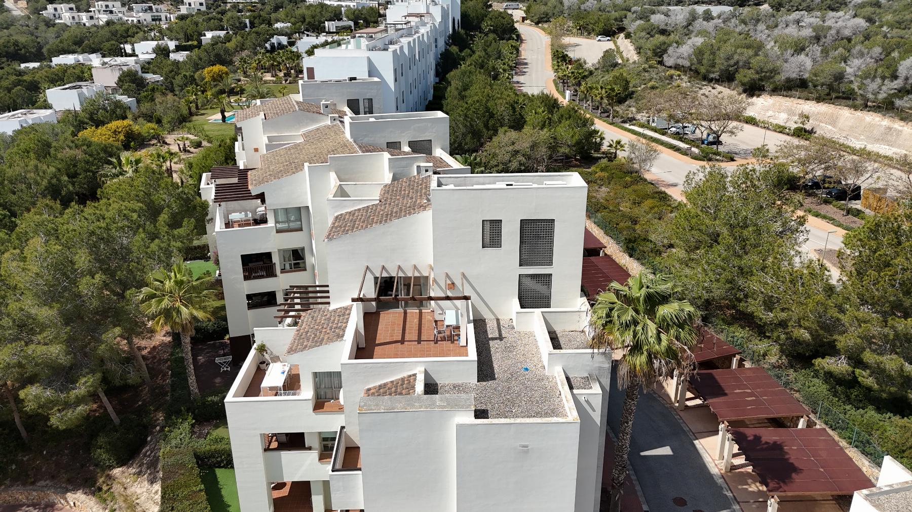 Aerial view of a white modern apartment complex with terracotta-tiled rooftops set among trees and a curving road nearby.
