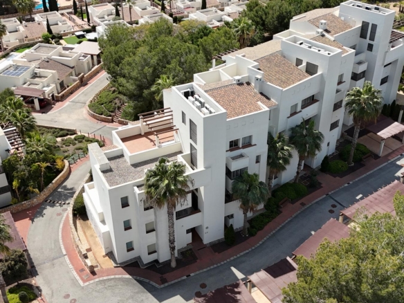 Aerial view of a white, multi-story residential complex with terracotta roofs, palm trees, and red-paved streets in a sunny area.