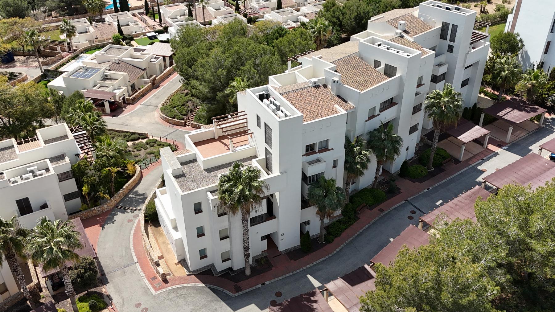 Aerial view of a white, multi-story residential complex with terracotta roofs, palm trees, and red-paved streets in a sunny area.