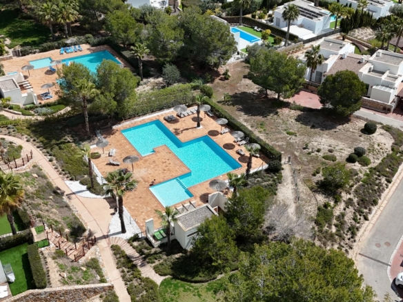 Aerial view of a resort complex with two turquoise swimming pools surrounded by terracotta decks, sun loungers, palm trees, and white villa-style buildings nearby.