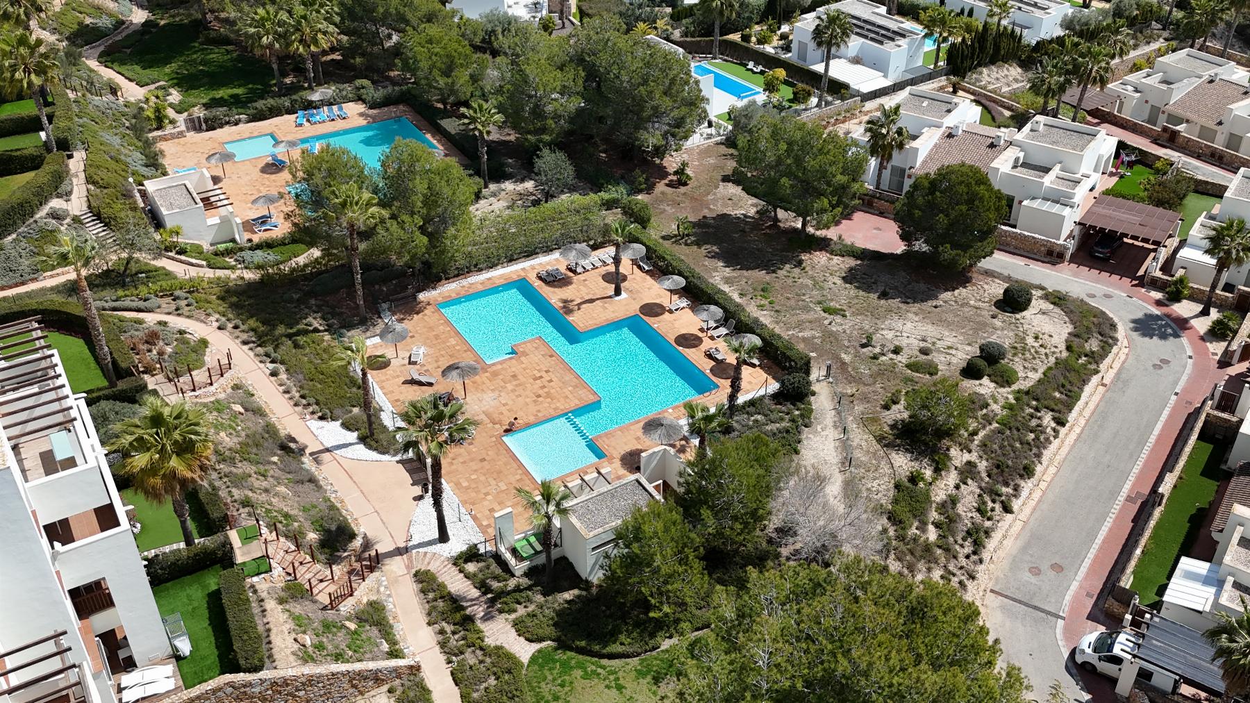 Aerial view of a resort complex with two turquoise swimming pools surrounded by terracotta decks, sun loungers, palm trees, and white villa-style buildings nearby.