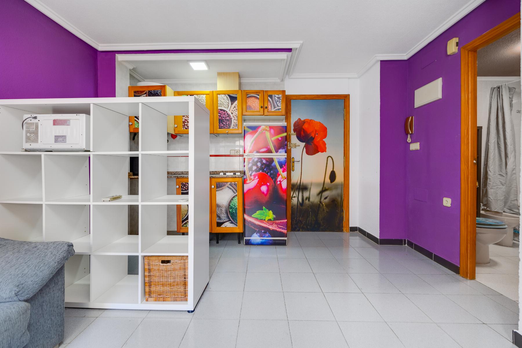 Bright interior with purple accent walls, white open shelving, and a back kitchen with orange cabinets and a floral-fruit refrigerator panel.