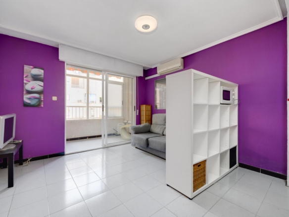 Living room with purple accent wall, gray sofa, white cubby shelf, microwave on top, and sliding glass doors to a balcony visible outside.