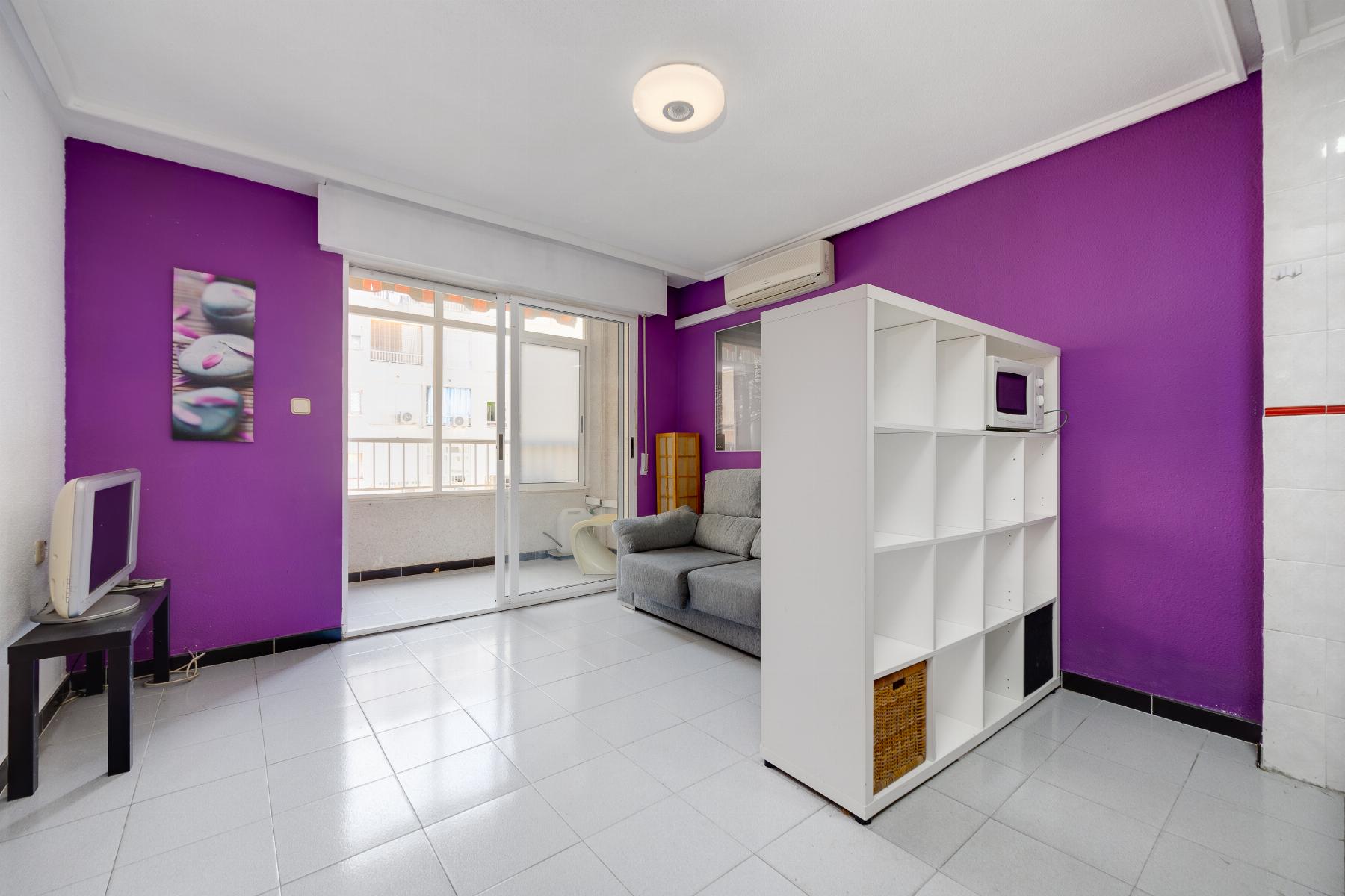 Living room with purple accent wall, gray sofa, white cubby shelf, microwave on top, and sliding glass doors to a balcony visible outside.