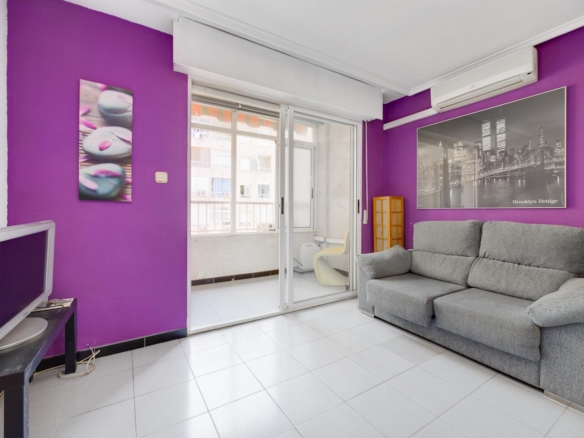 Living room with purple accent wall, gray sectional sofa, Brooklyn Bridge artwork, balcony through sliding glass doors, and an old computer monitor on a desk.