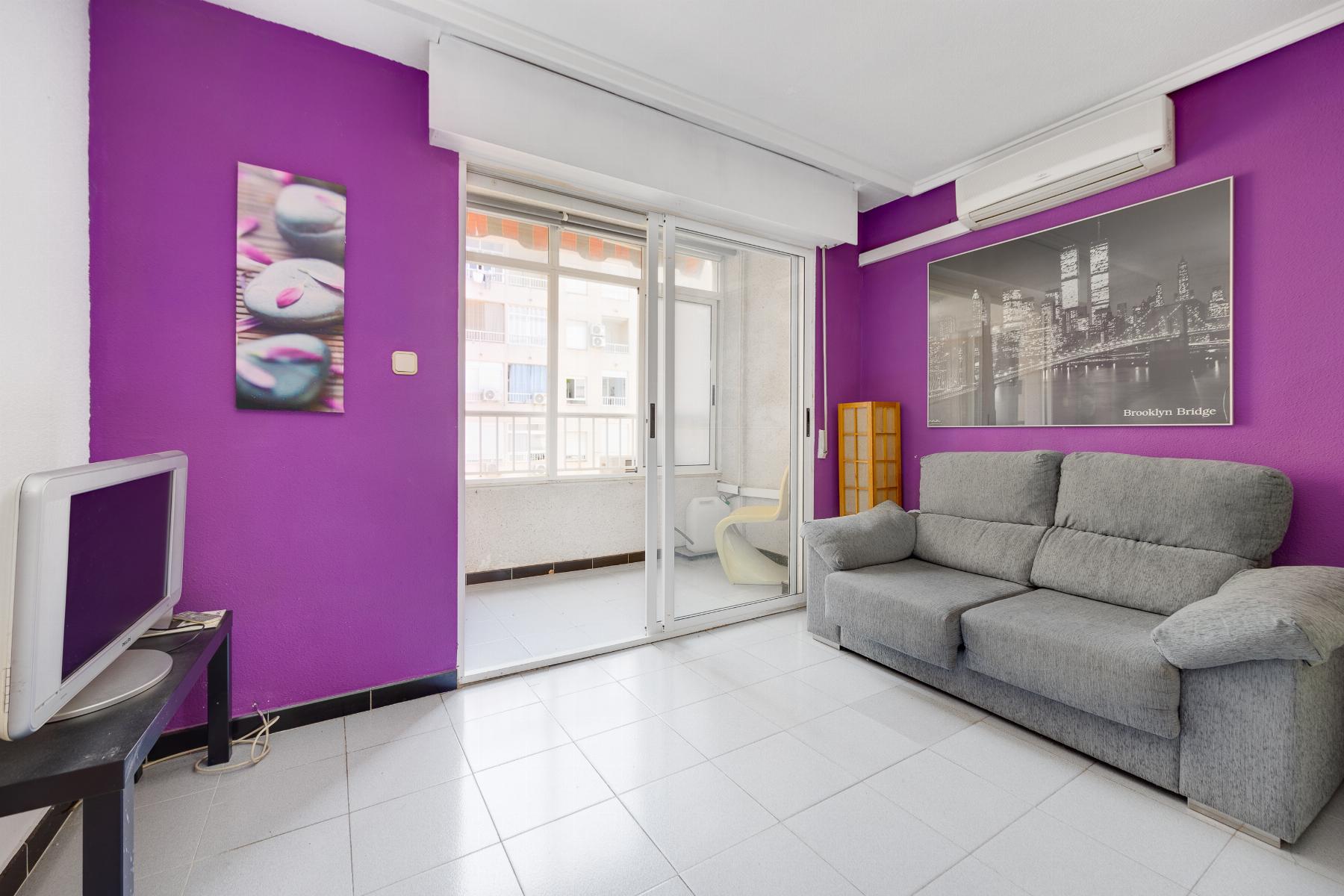 Living room with purple accent wall, gray sectional sofa, Brooklyn Bridge artwork, balcony through sliding glass doors, and an old computer monitor on a desk.