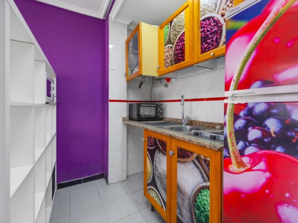 Compact kitchen with purple wall, white shelves, microwave, granite counter, orange-framed cabinets with fruit photos, and a colorful fruit fridge.