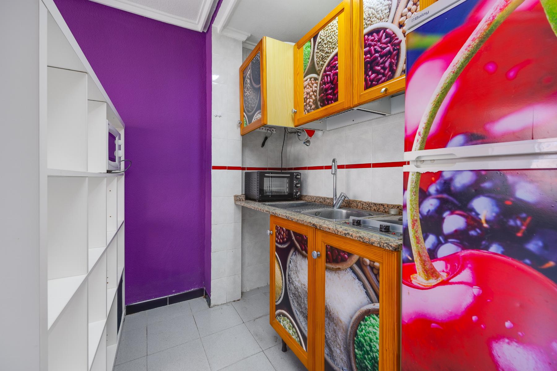 Compact kitchen with purple wall, white shelves, microwave, granite counter, orange-framed cabinets with fruit photos, and a colorful fruit fridge.