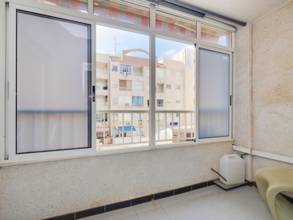 Enclosed balcony with sliding glass windows and a view of a beige apartment building across the way; foreground shows light tile floor, a white container, and pale yellow chairs at the right edge.