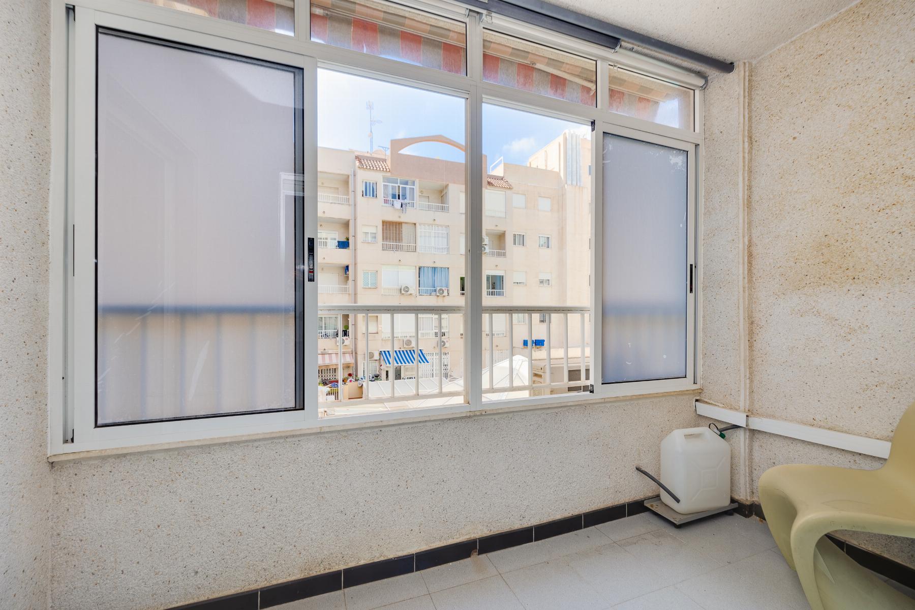 Enclosed balcony with sliding glass windows and a view of a beige apartment building across the way; foreground shows light tile floor, a white container, and pale yellow chairs at the right edge.