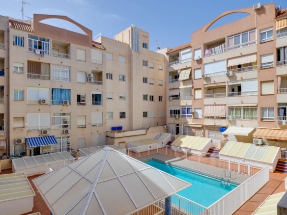 Courtyard of beige apartment buildings with balconies, air conditioners, and striped awnings surrounding a glass-roofed pool area on a sunny day.
