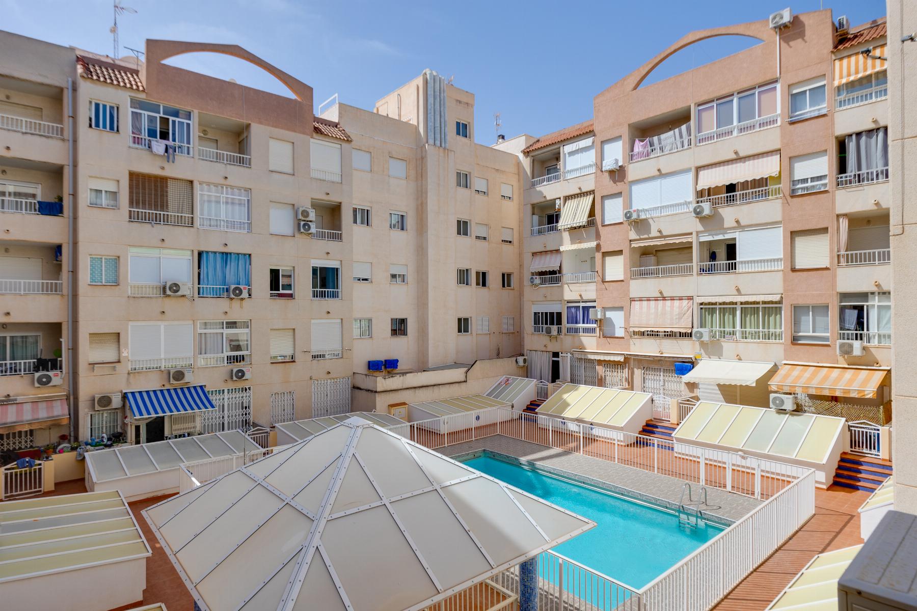 Courtyard of beige apartment buildings with balconies, air conditioners, and striped awnings surrounding a glass-roofed pool area on a sunny day.