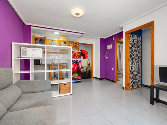Living room with a purple accent wall, gray sectional sofa, and a white modular shelf unit near the kitchen area behind it.
