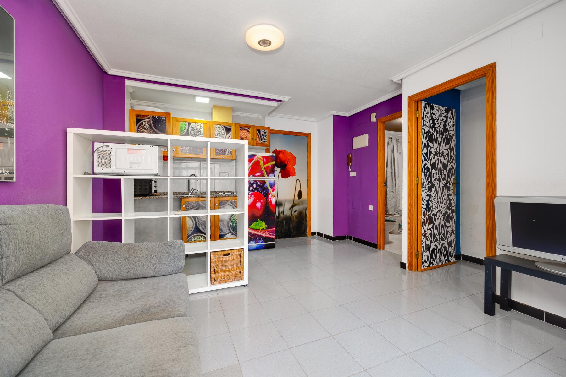 Living room with a purple accent wall, gray sectional sofa, and a white modular shelf unit near the kitchen area behind it.