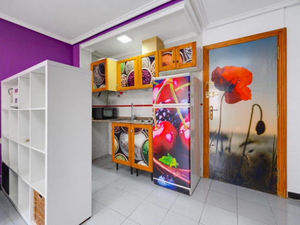 Colorful kitchen interior with a purple wall, white shelving, wooden cabinets with fruit-pattern doors, and a fridge covered in fruit imagery.