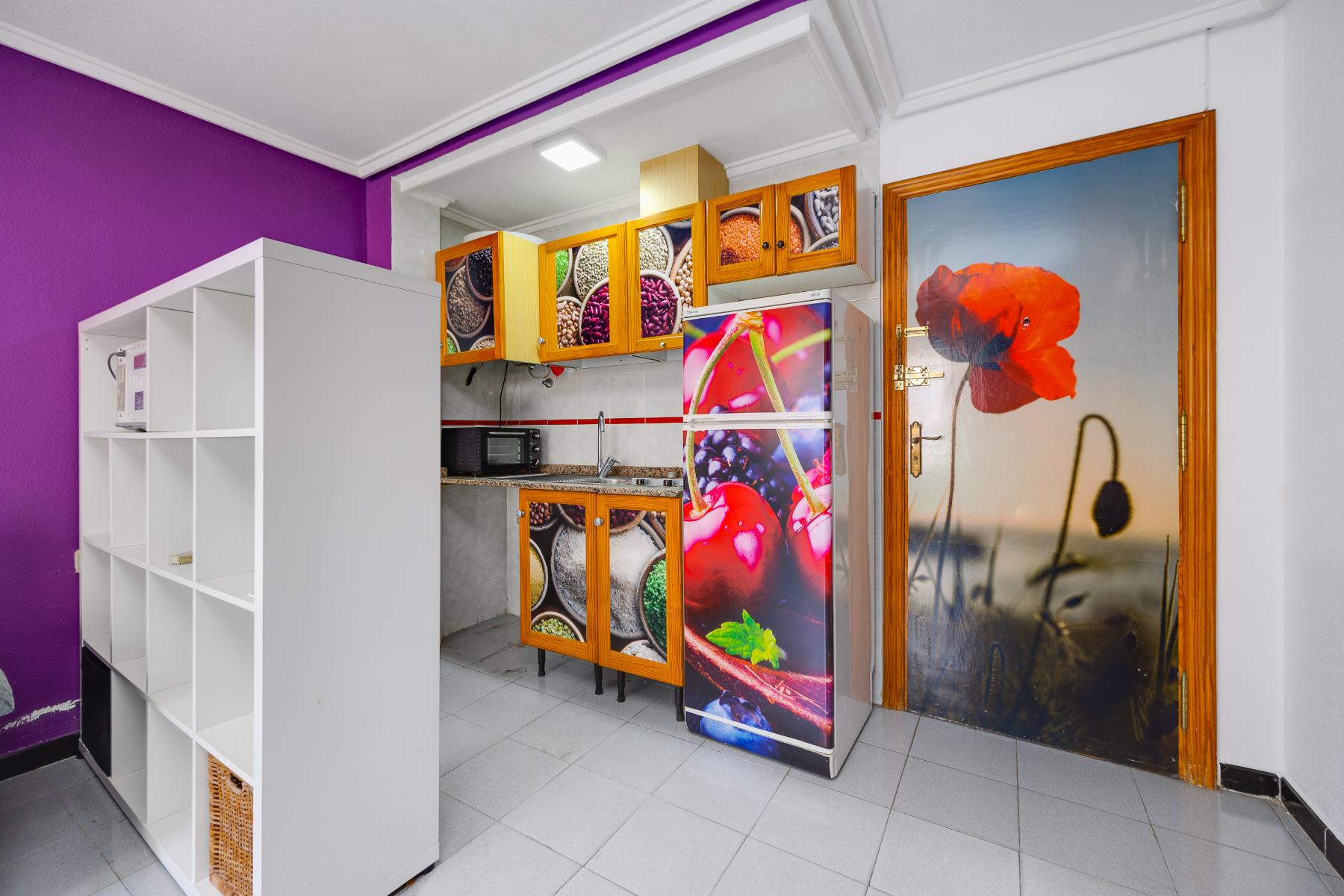 Colorful kitchen interior with a purple wall, white shelving, wooden cabinets with fruit-pattern doors, and a fridge covered in fruit imagery.