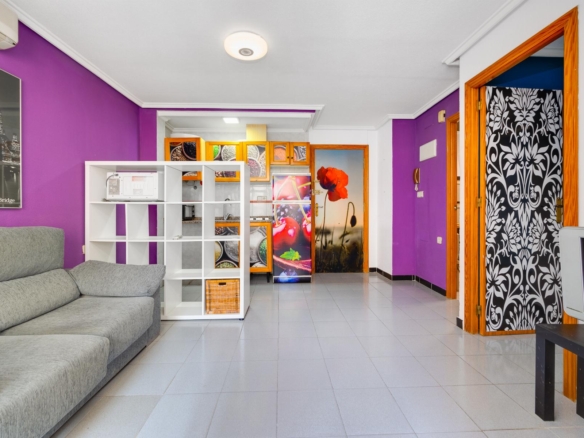 Living room with purple walls, gray sectional sofa, and white shelving unit near a colorful kitchen area with wooden cabinets.