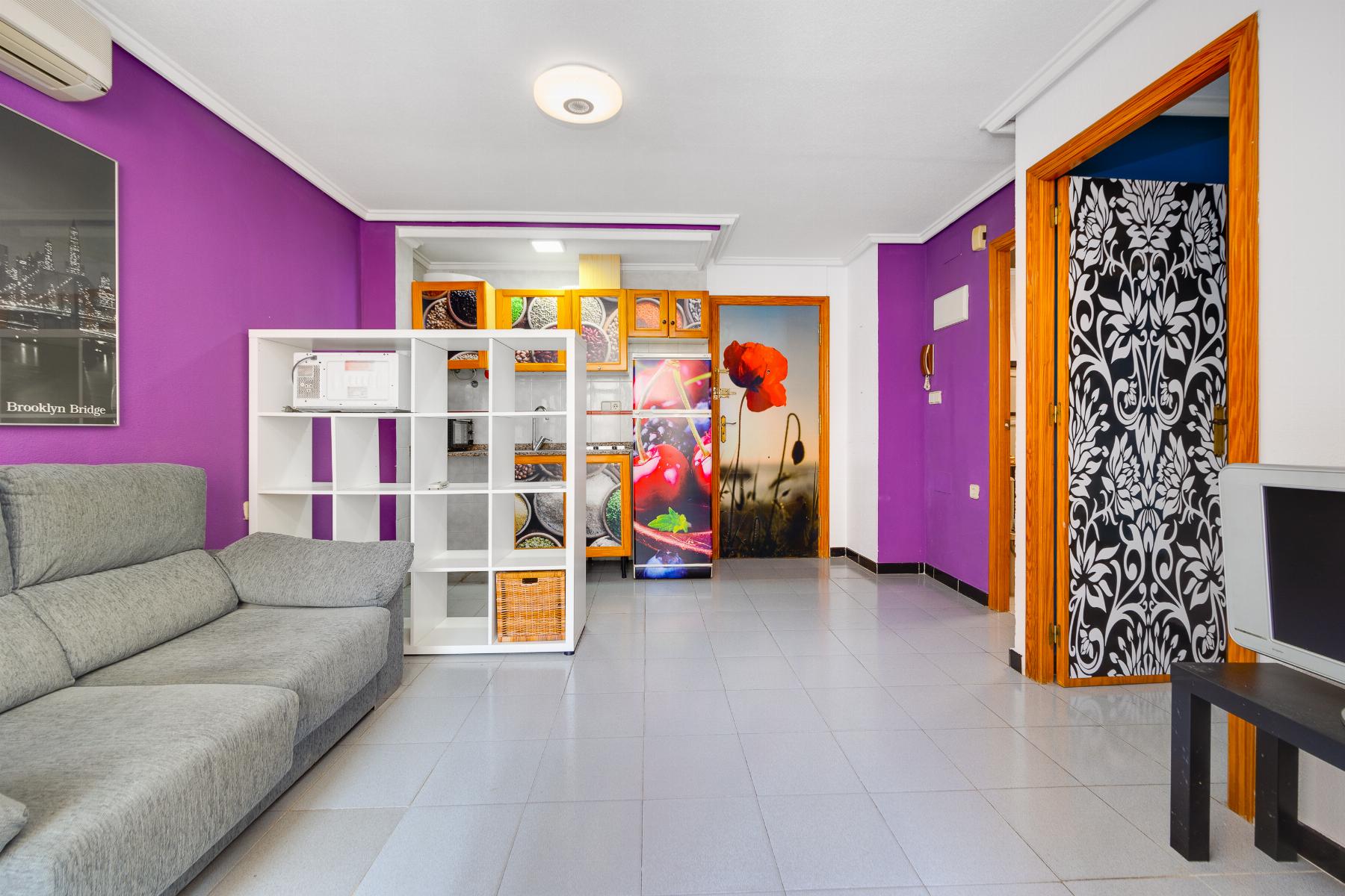 Living room with purple walls, gray sectional sofa, and white shelving unit near a colorful kitchen area with wooden cabinets.