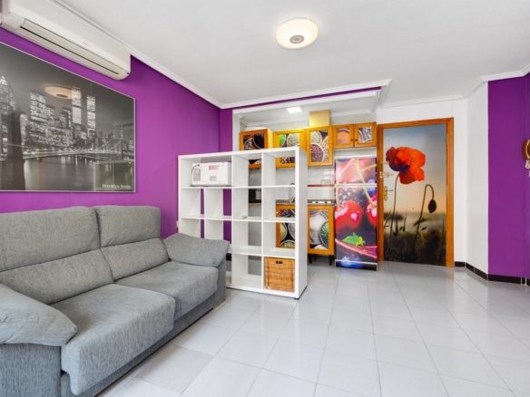 Living room with purple accent walls, a gray sectional sofa, and a white cube storage unit by the kitchen area. A door features a large red flower image.
