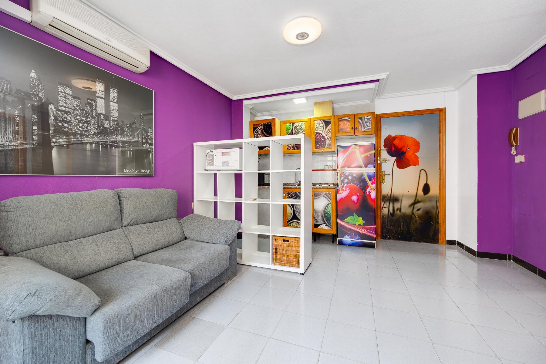 Living room with purple accent walls, a gray sectional sofa, and a white cube storage unit by the kitchen area. A door features a large red flower image.