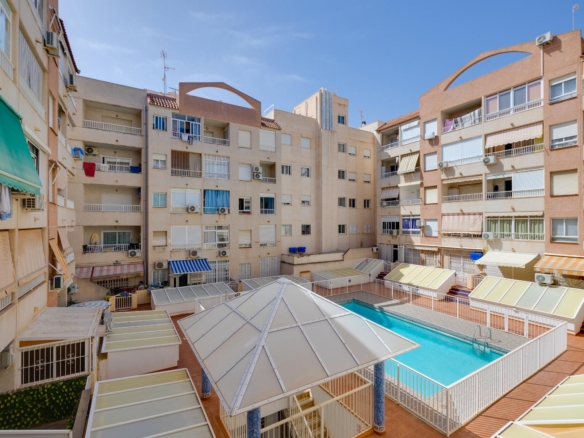 Courtyard of beige apartment buildings with balconies surrounding a rectangular blue pool and glass-roofed walkways beneath a clear sky