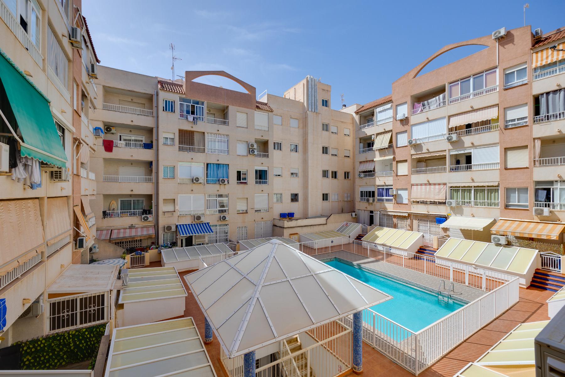 Courtyard of beige apartment buildings with balconies surrounding a rectangular blue pool and glass-roofed walkways beneath a clear sky