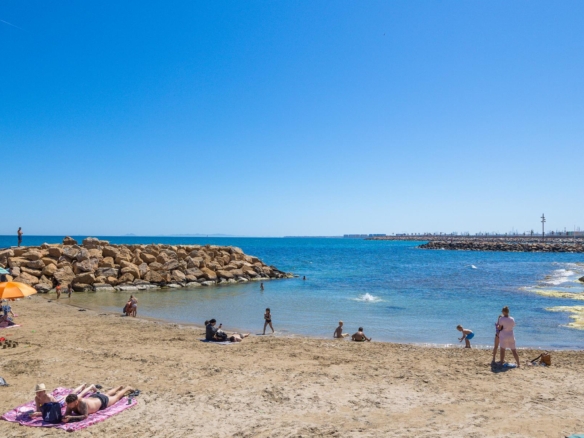 Sunny beach scene with people sunbathing on towels, an orange umbrella, and a rocky breakwater calm blue sea.