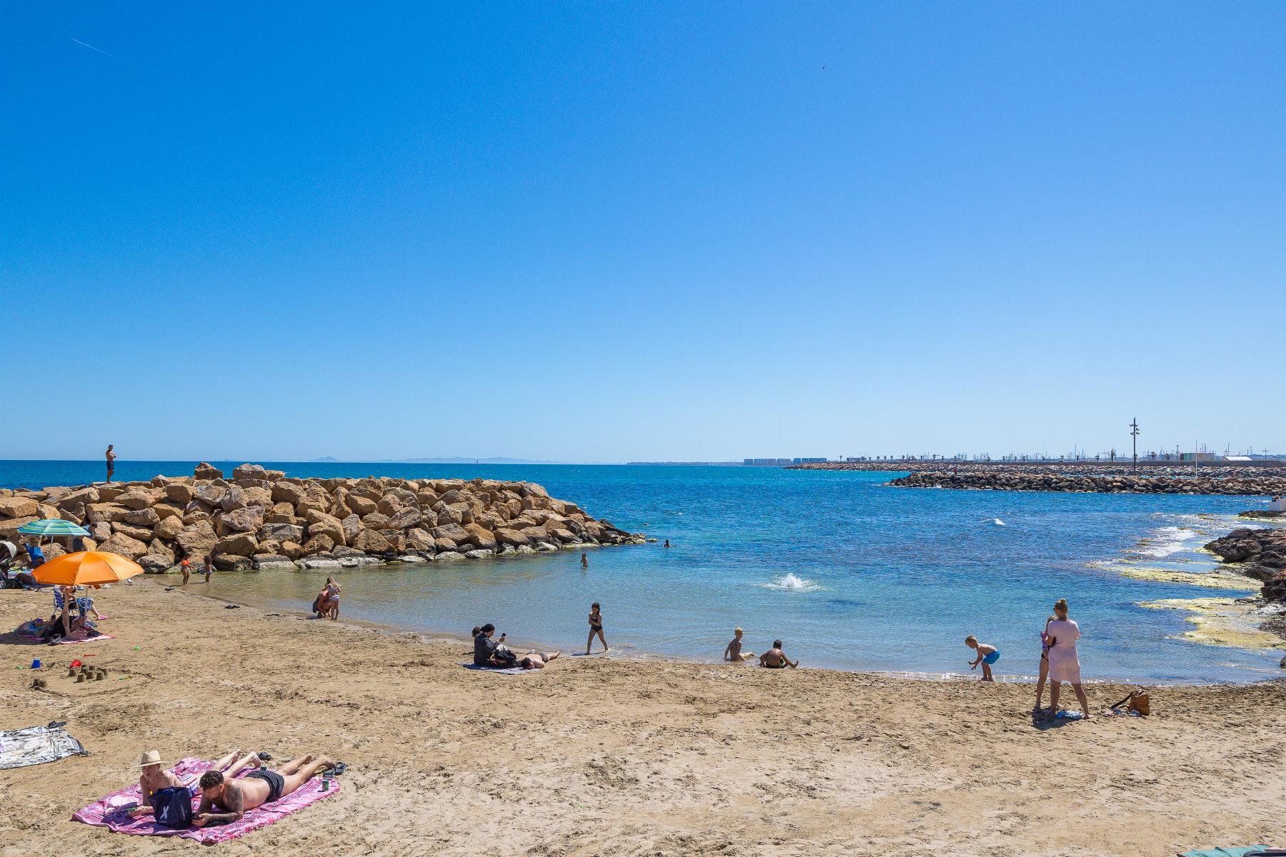 Sunny beach scene with people sunbathing on towels, an orange umbrella, and a rocky breakwater calm blue sea.