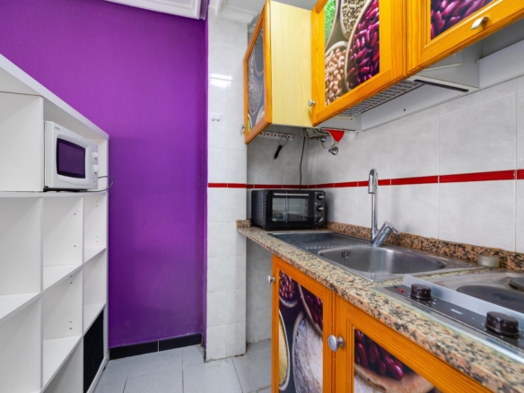 Small kitchen with a bright purple wall, white open shelving, and a countertop with a toaster oven and sink visible along tiled backsplash and red stripe accent tiles.