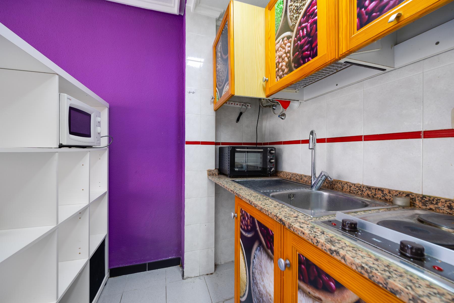 Small kitchen with a bright purple wall, white open shelving, and a countertop with a toaster oven and sink visible along tiled backsplash and red stripe accent tiles.