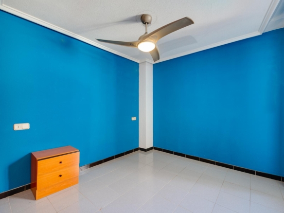 Blue corner room with white ceiling fan, small wooden dresser, and light gray tiled floor with black baseboard trim.