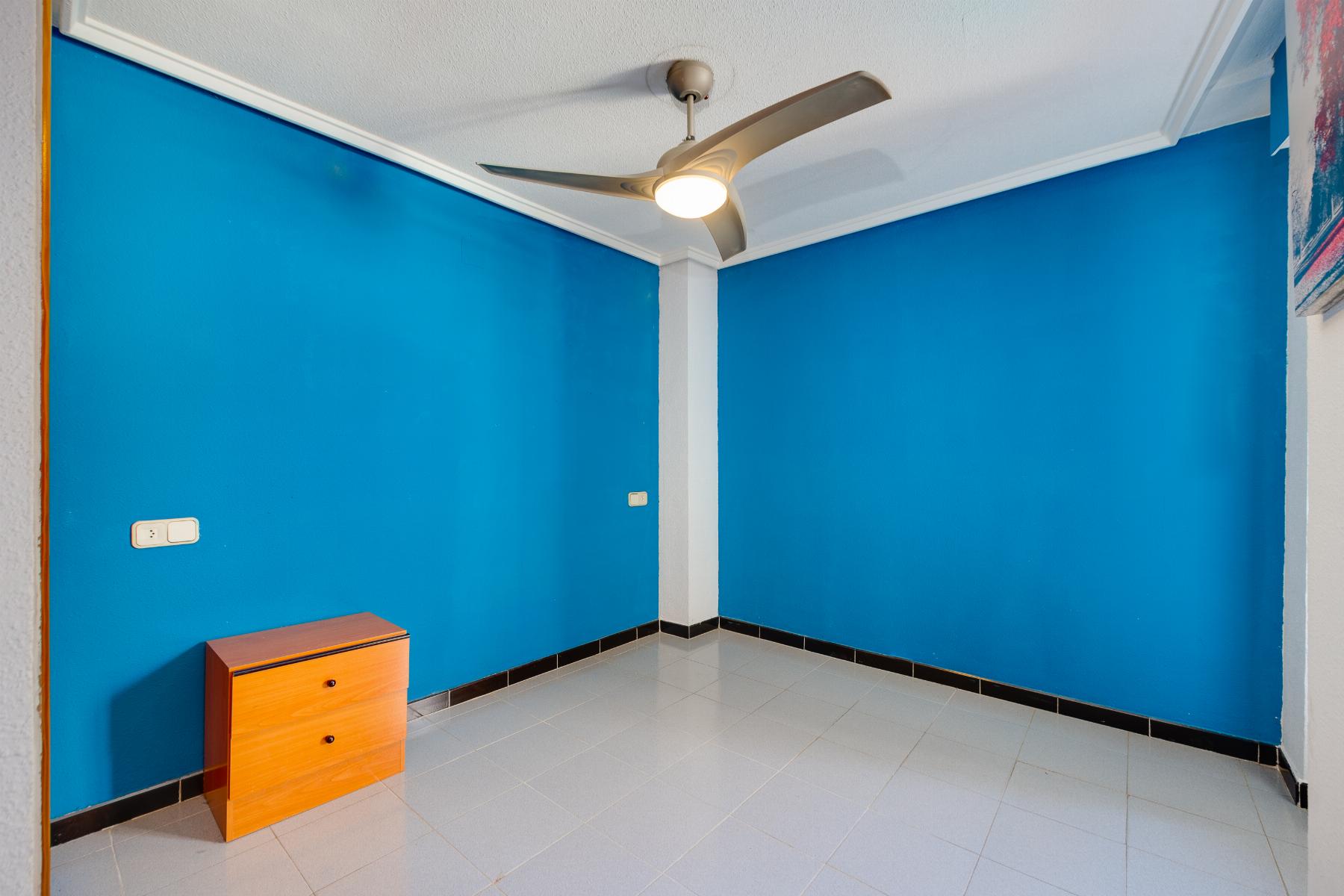 Blue corner room with white ceiling fan, small wooden dresser, and light gray tiled floor with black baseboard trim.