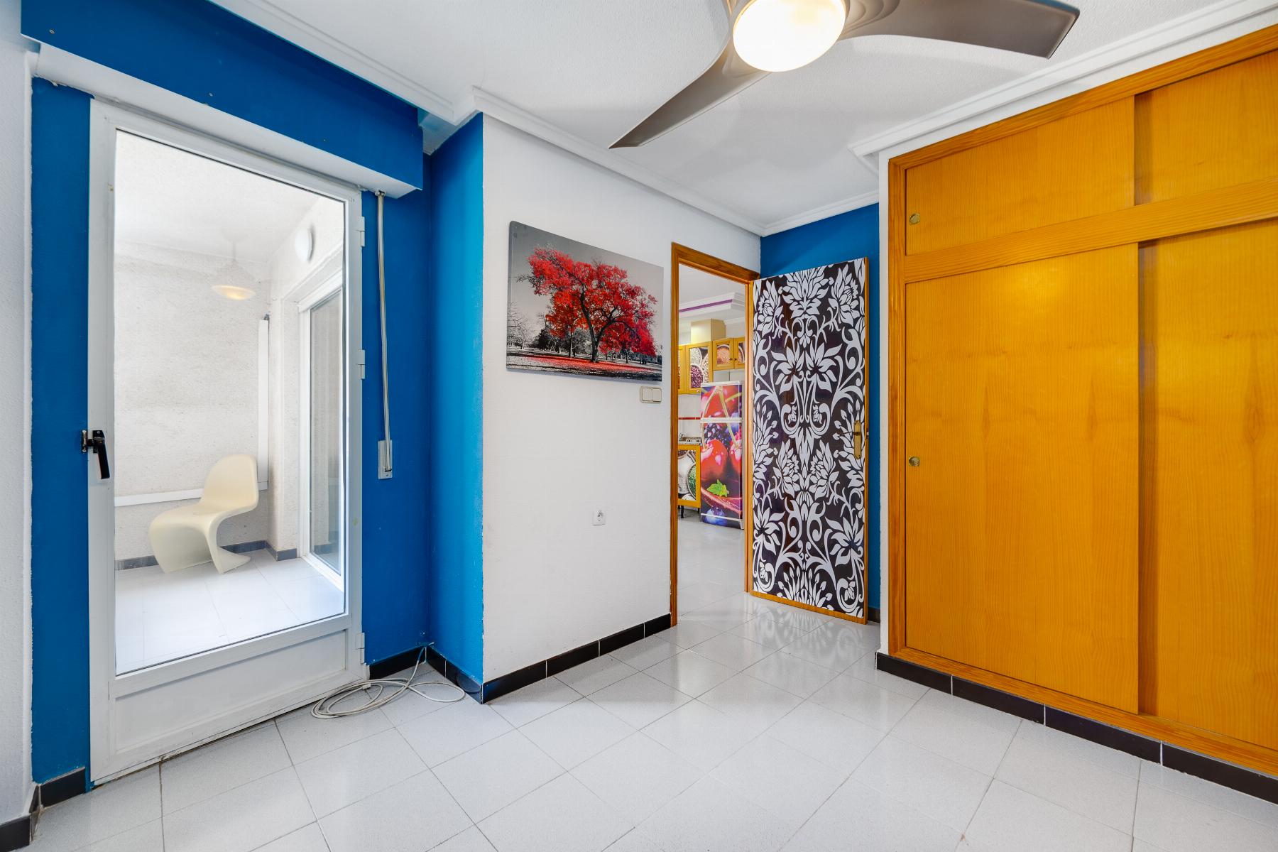 Interior hallway with blue walls, white trim, and a large wooden closet on the right. A framed red-tree artwork hangs on the wall; a patterned screen and a doorway lead to another room with colorful decor visible.