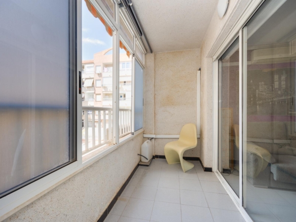 Enclosed apartment balcony with a beige curved chair, tiled floor, and sliding glass doors; neighboring buildings visible outside the window.