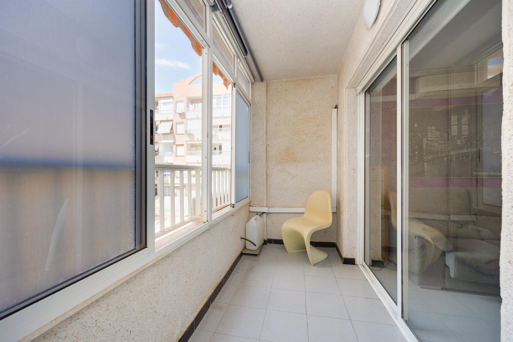 Enclosed apartment balcony with a beige curved chair, tiled floor, and sliding glass doors; neighboring buildings visible outside the window.