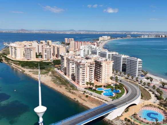 Aerial view of a coastal city with beige and white apartment blocks lining a curved shoreline, turquoise water, and a distant horizon of low hills under a blue sky. A highway bridge arcs over a waterway in the foreground, with a circular palm-filled pool area and palm trees nearby.