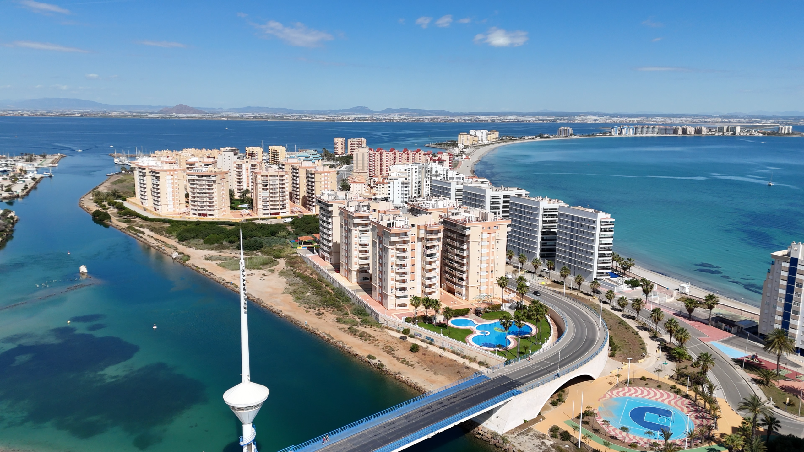 Aerial view of a coastal city with beige and white apartment blocks lining a curved shoreline, turquoise water, and a distant horizon of low hills under a blue sky. A highway bridge arcs over a waterway in the foreground, with a circular palm-filled pool area and palm trees nearby.