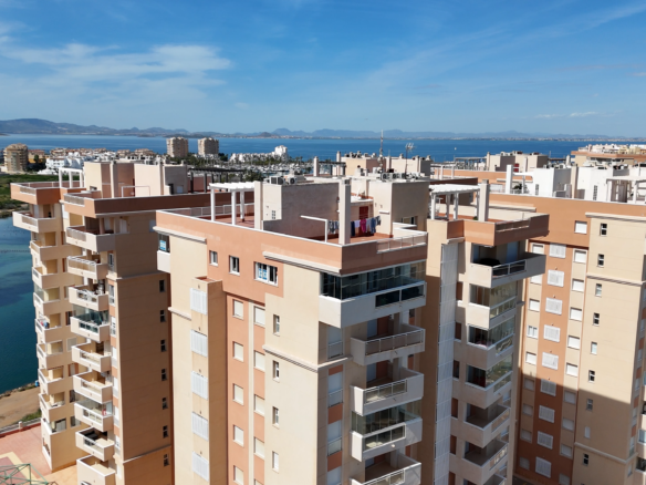 Cluster of beige apartment buildings along a waterway with a calm blue sea and distant mountains under a clear sky.