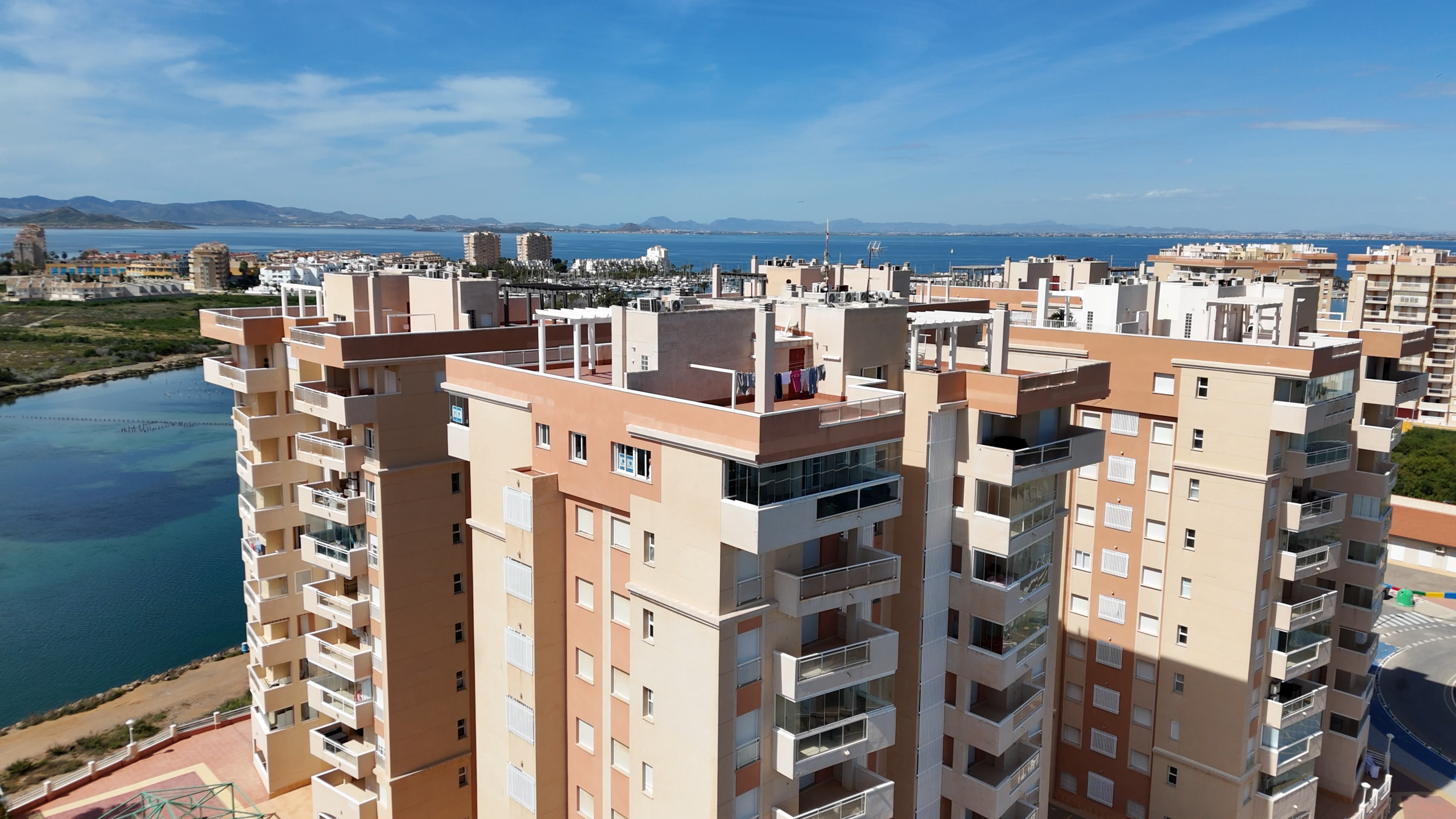 Cluster of beige apartment buildings along a waterway with a calm blue sea and distant mountains under a clear sky.