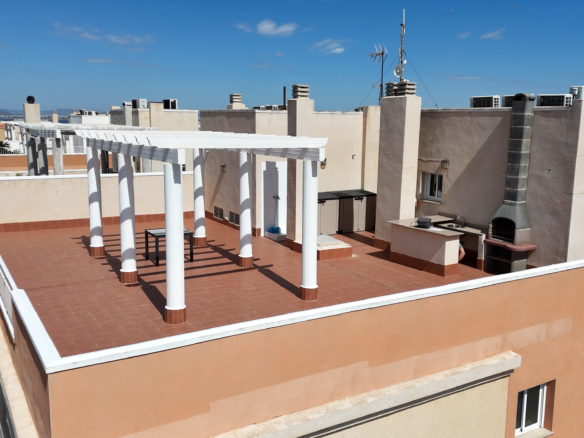 Rooftop terrace with white pergola columns, a built-in barbecue, and terracotta tiles, overlooking city buildings and the sea under a blue sky.