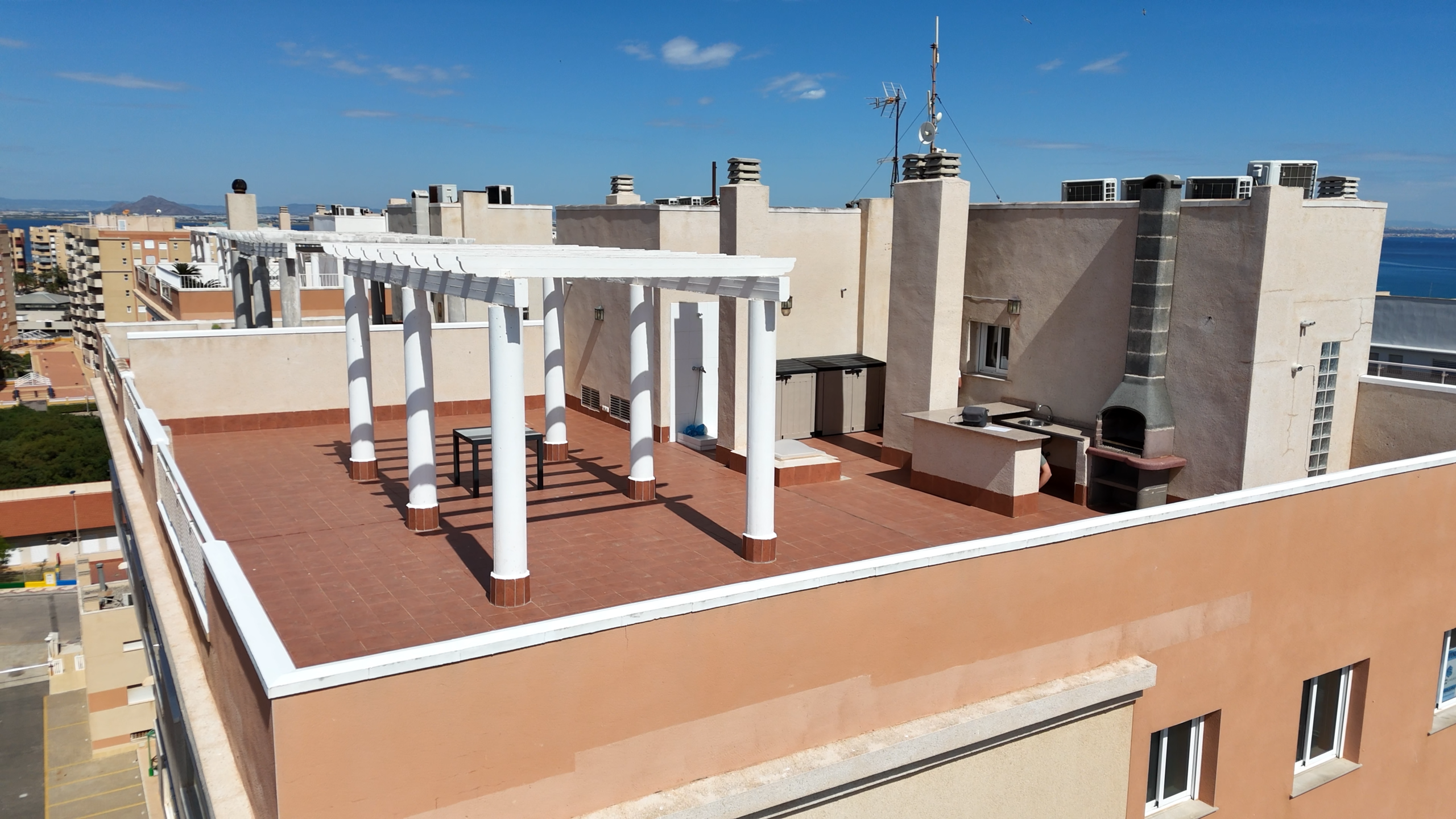 Rooftop terrace with white pergola columns, a built-in barbecue, and terracotta tiles, overlooking city buildings and the sea under a blue sky.
