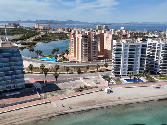 Aerial view of a coastal city with tall white and beige apartment towers, a marina, and distant mountains across the bay.