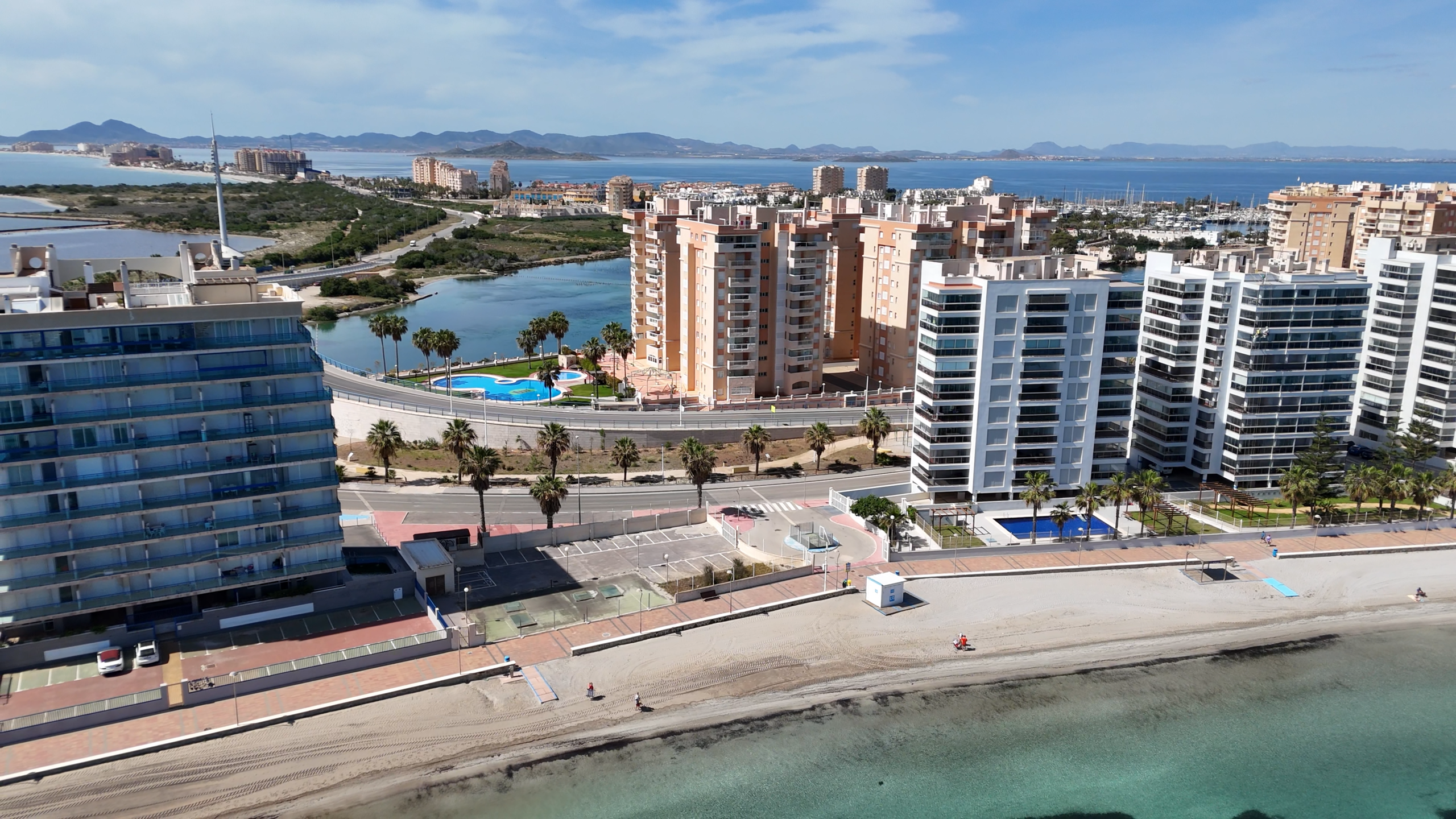 Aerial view of a coastal city with tall white and beige apartment towers, a marina, and distant mountains across the bay.